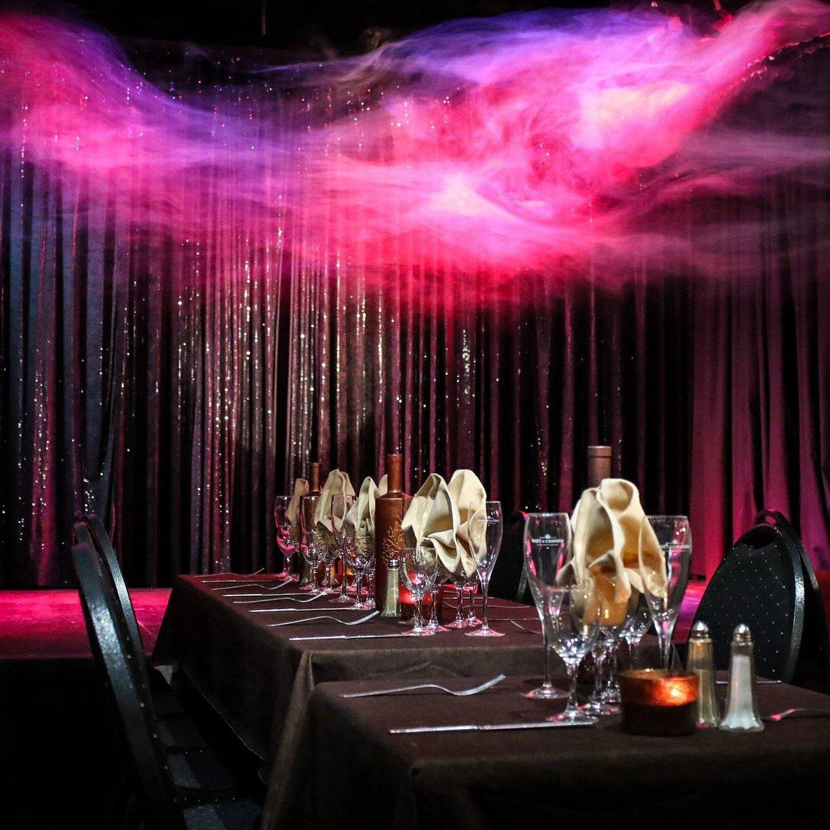 Table dressée au cabaret avec verres, couverts et serviettes pliées, devant des rideaux éclairés en rose et violet, sans artiste sur scène.